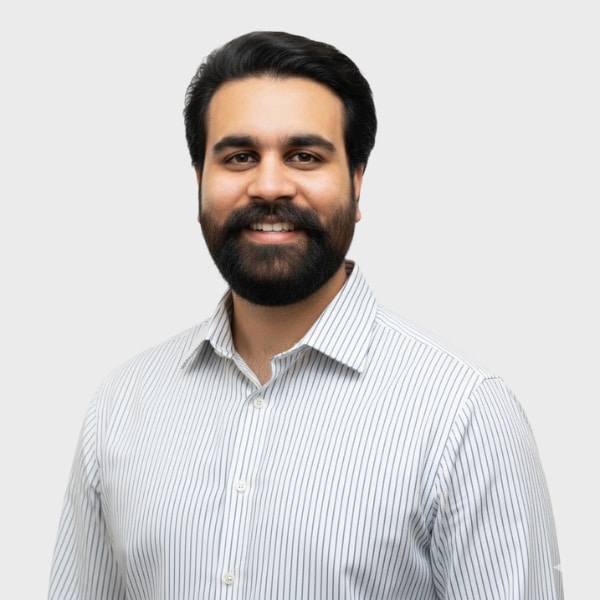 A man with dark hair and a beard, wearing a white shirt with thin vertical stripes, smiles confidently as he represents an IT staffing agency in Toronto against a plain light gray background.