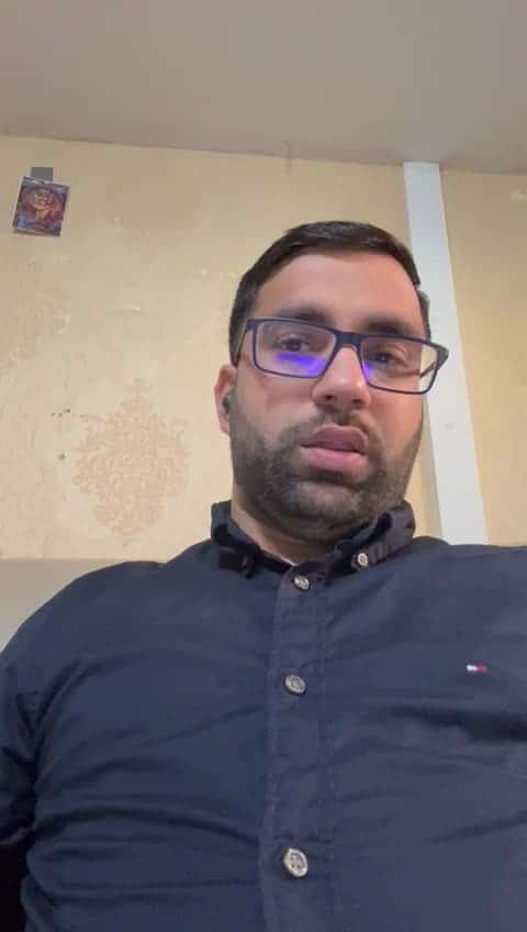 A man with short dark hair, a beard, and glasses is wearing a navy blue button-up shirt and sitting indoors against a beige wall—an ideal look for someone seeking IT and Finance jobs in Toronto.