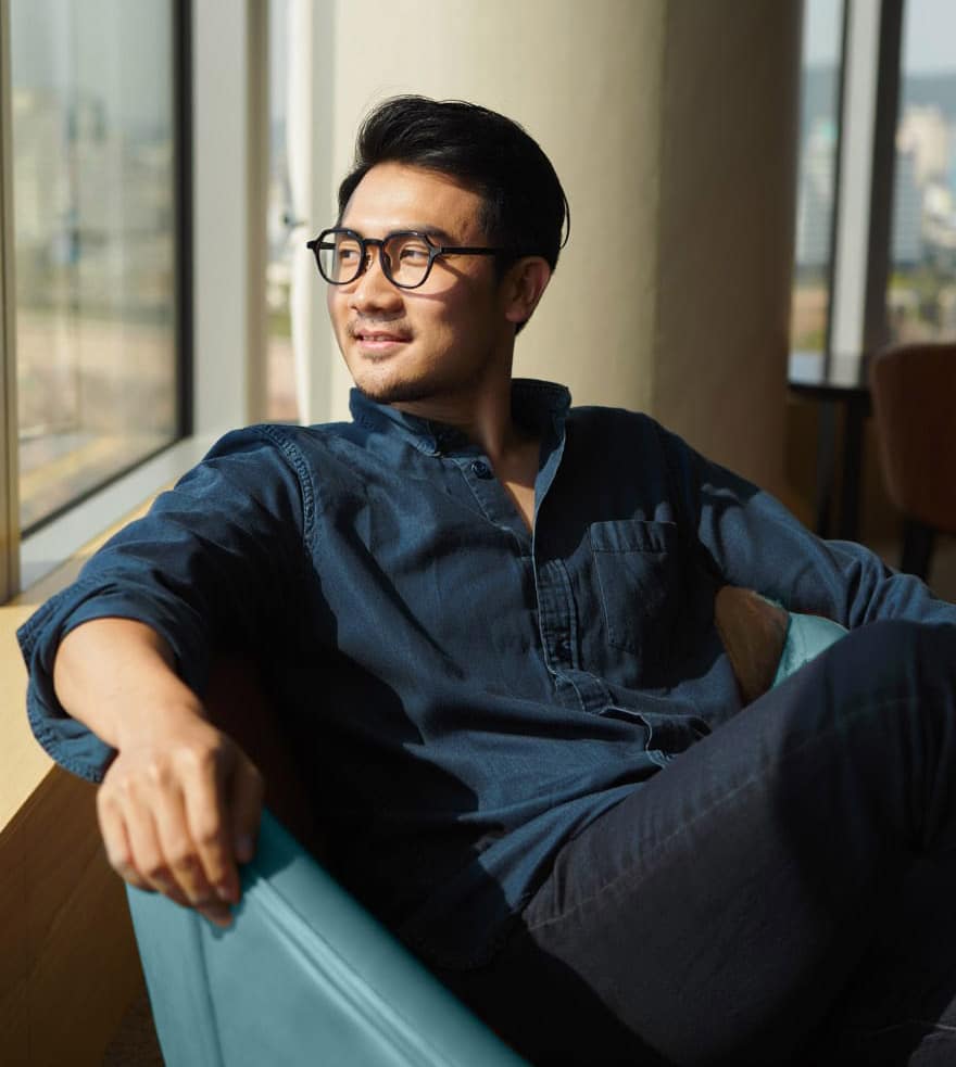 A man wearing glasses and a dark blue shirt sits in a chair by a large window, smiling and looking outside at the sunlight, reflecting on the opportunities that Permanent and Contract Staffing Services can bring to his career.