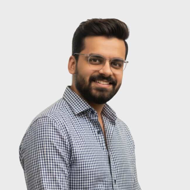 A man with glasses, a beard, and short dark hair smiles while wearing a black-and-white checkered shirt, standing against a plain white background, representing an IT staffing agency in Toronto.