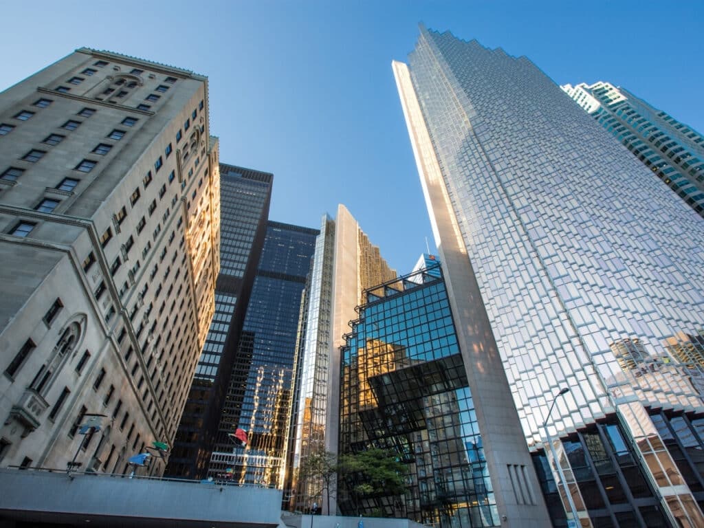 Tall modern and historic office buildings rise against a clear blue sky in a cityscape, reflecting sunlight and each other in their glass facades, with a hint of trees and urban street at the bottom.